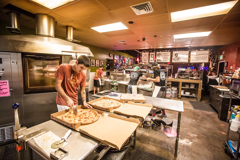 A man cuts a pizza at Hot Tomato while other employees put toppings on the pizzas in the background.