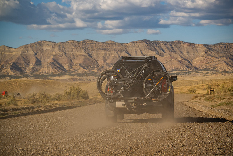 A black car with a bike on the back drives on a dirt road.