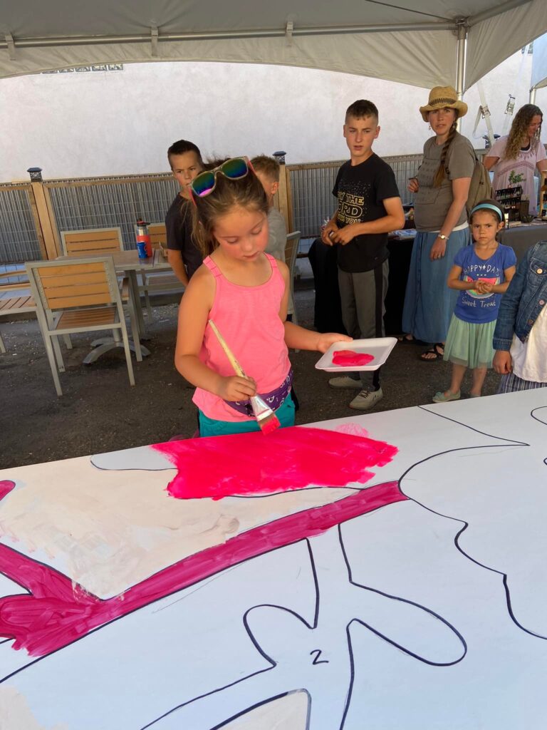 A young girl paints on a poster while people watch in the background.
